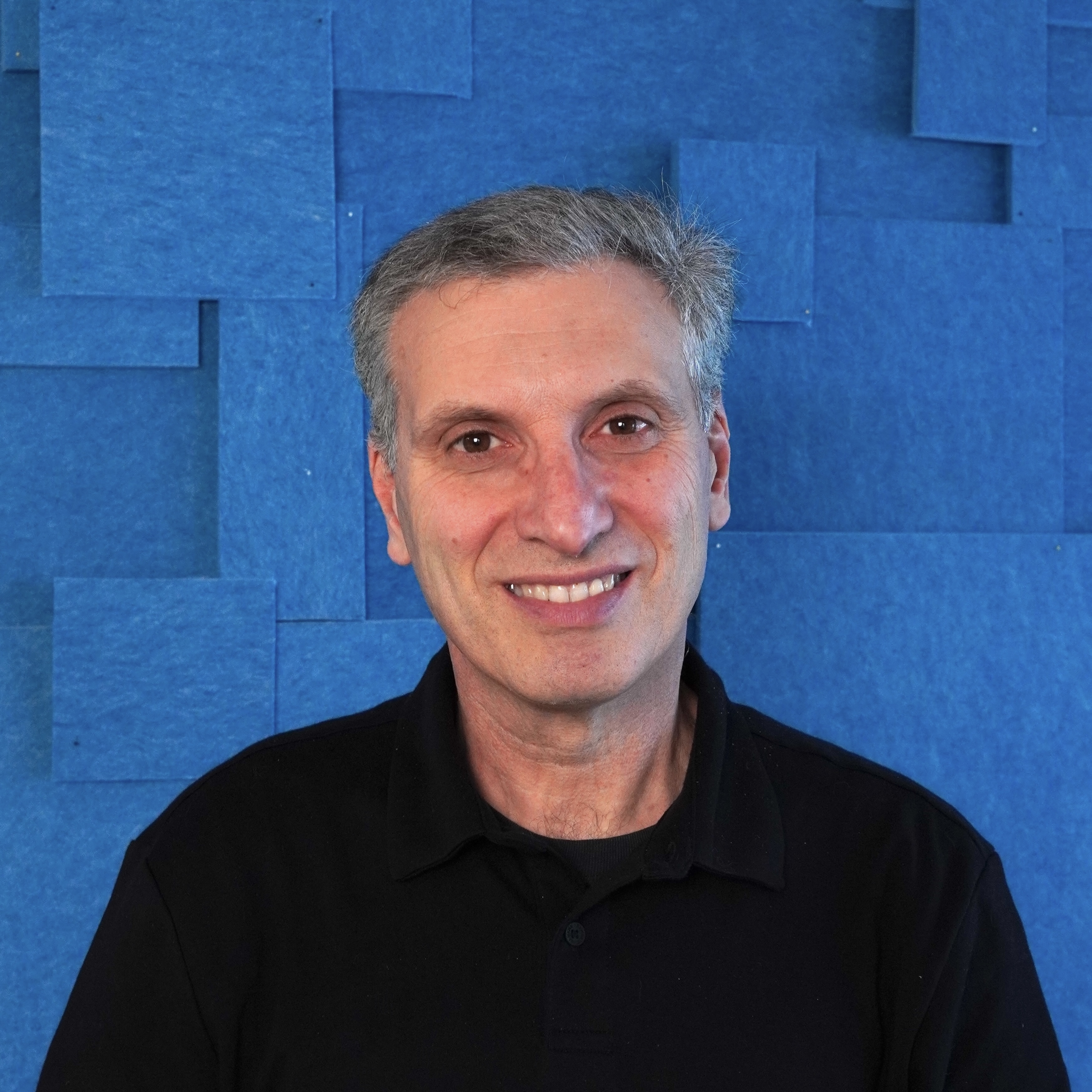 Dark-haired man standing in front of a textured blue background.