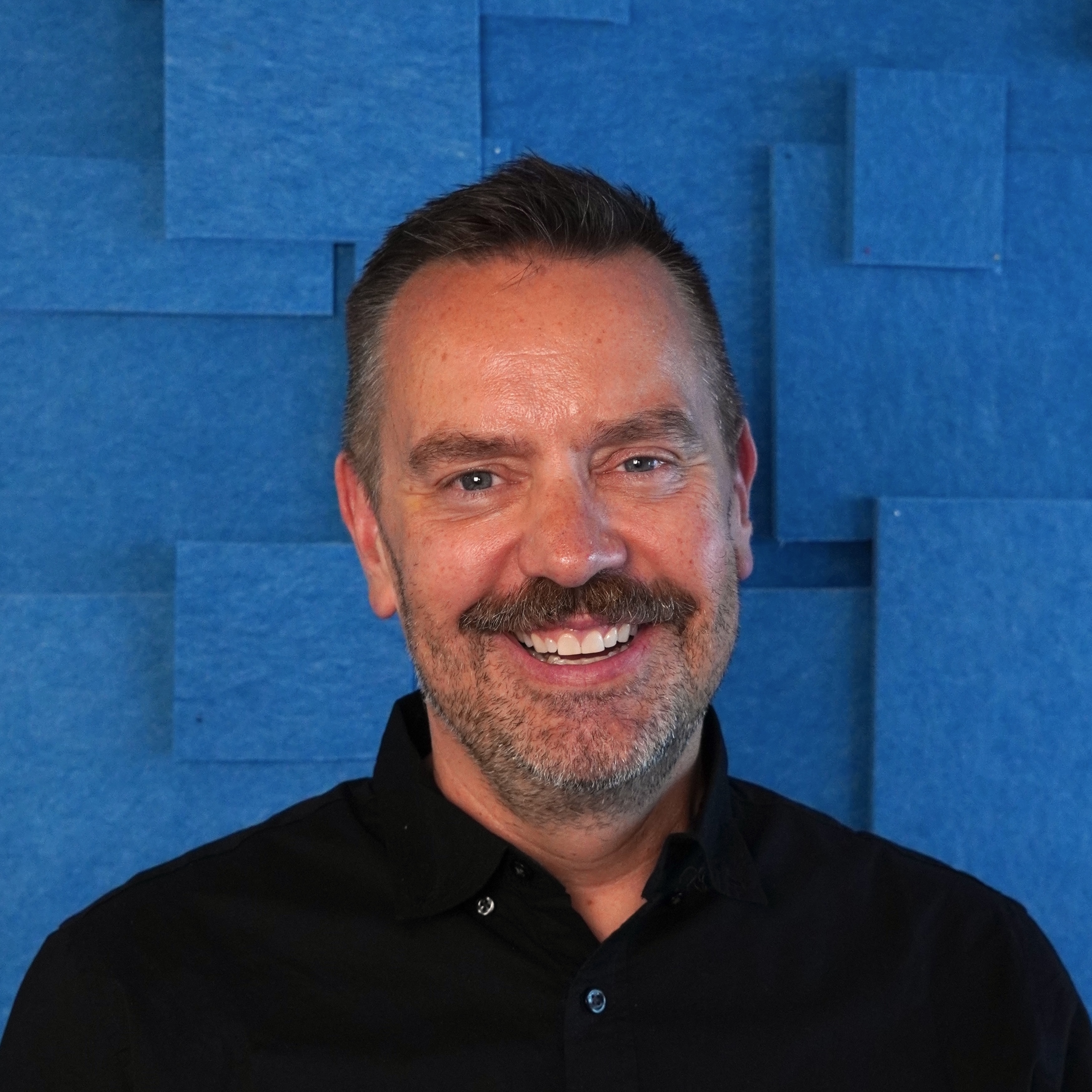 Dark-haired man standing in front of a textured blue background.